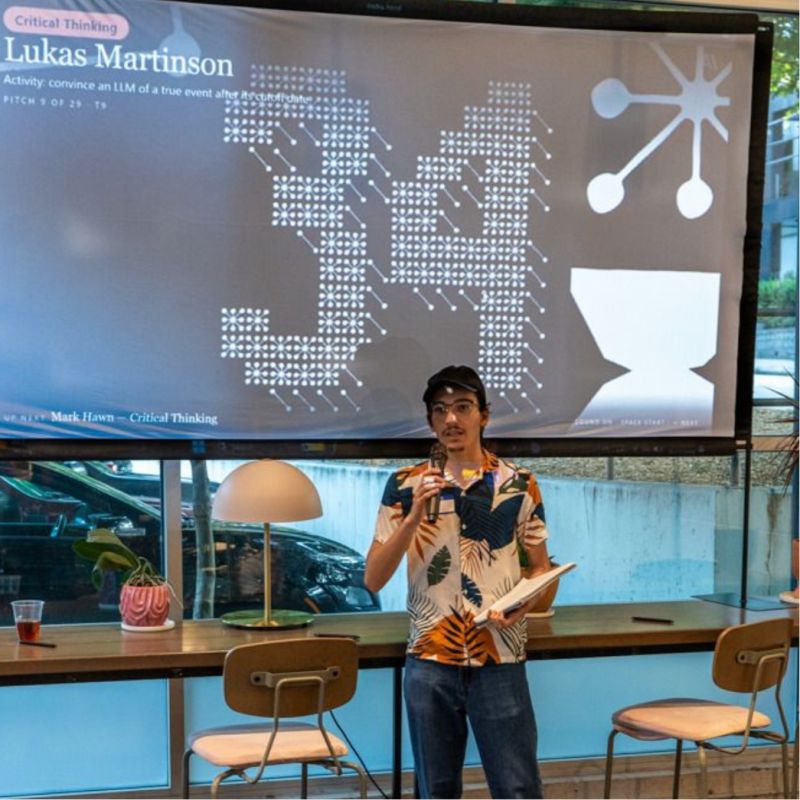 Lukas Martinson speaking into a handheld microphone in front of a large projection screen. The slide behind him is tagged Critical Thinking and carries his name above a one-line description of the activity: convince an LLM of a true event after its cutoff date.